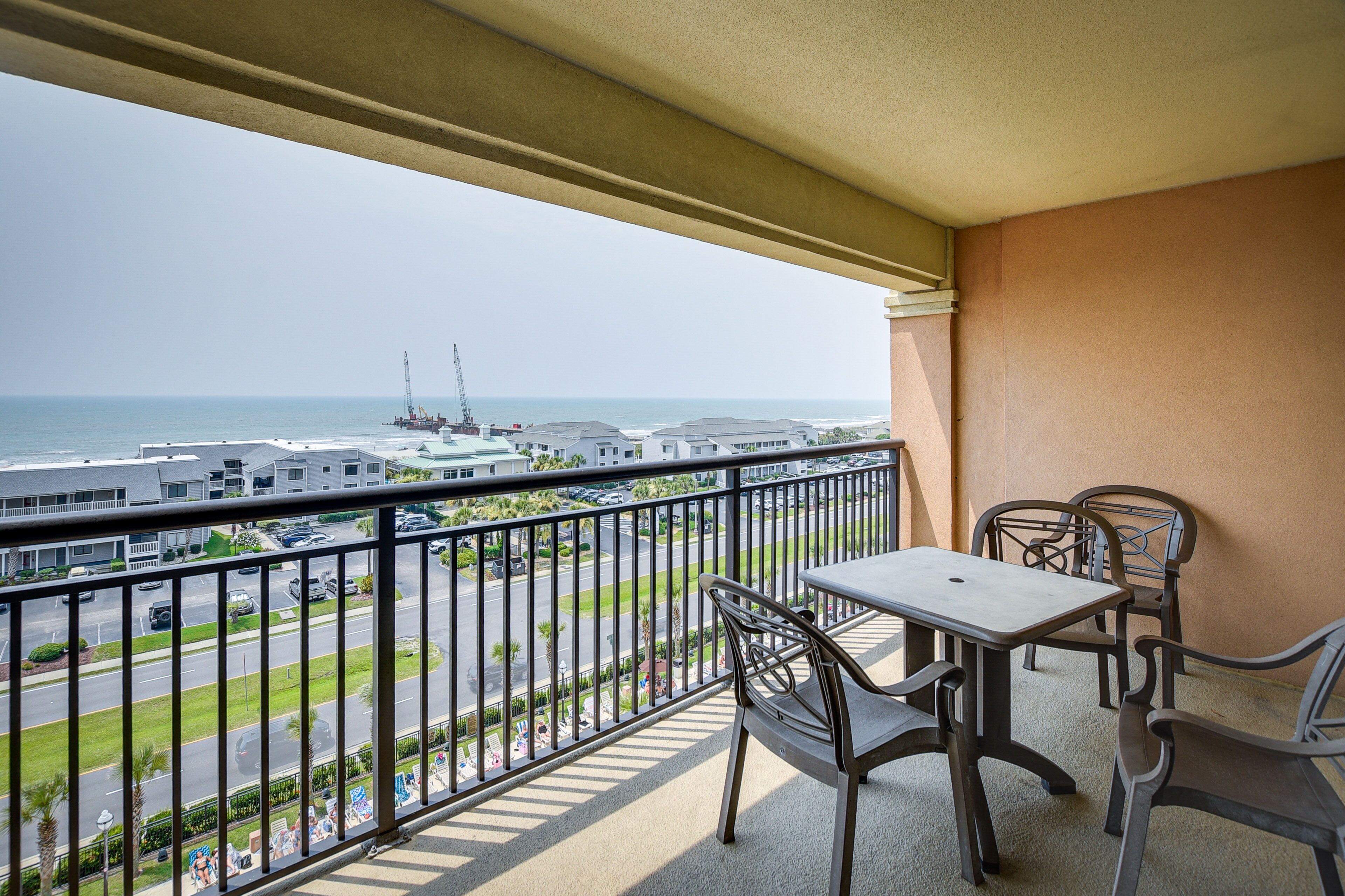 A balcony with a table and chairs, overlooking a road and a marina with boats and a crane.