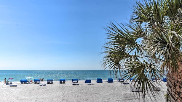 Une plage de sable blanc, sous un ciel bleu azur. Des chaises longues bleues et des parasols sont disposés le long du rivage.