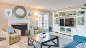 Un salon avec une cheminée, deux fauteuils, une table basse et un téléviseur posé sur un meuble.