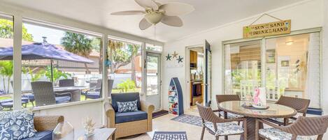A sunroom with a ceiling fan, wicker furniture, a glass table, and a sign reading "The Sun and the Sand Drink in the Hand".