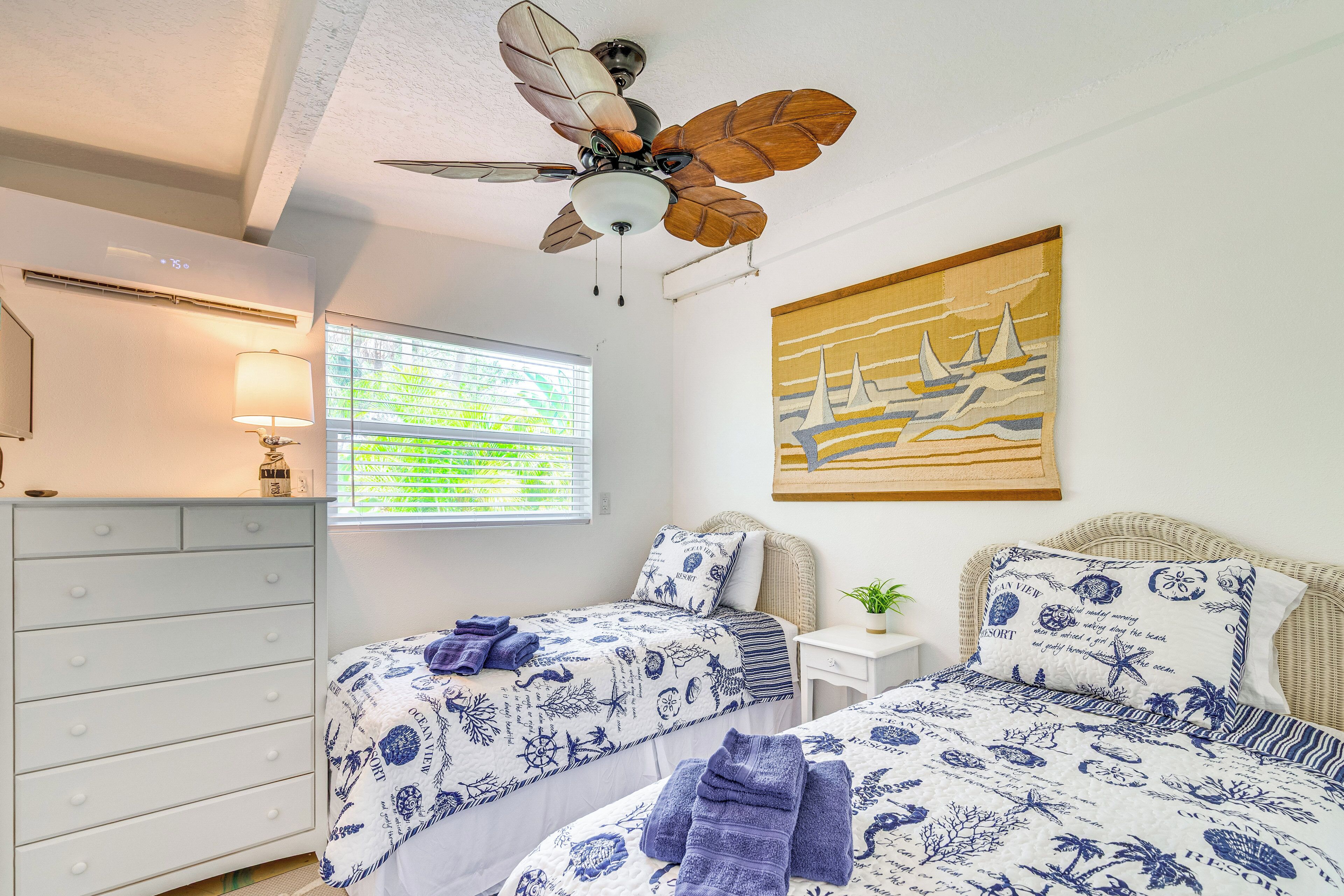 A bedroom with two beds, a ceiling fan, a window with blinds, a wall-mounted air conditioner, and a painting of sailboats.