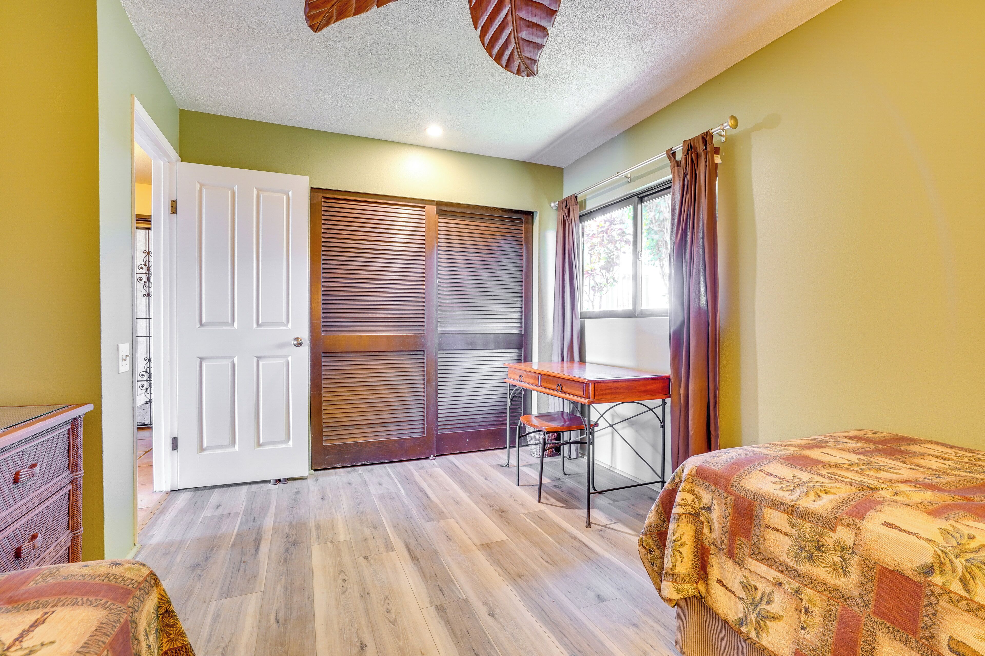 A room with a wooden floor, a desk with a chair, a bed with a patterned bedspread, a wardrobe, and a window with curtains.