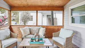 Une véranda couverte avec des meubles en osier, une table avec une bougie et une vue sur une maison et des arbres.