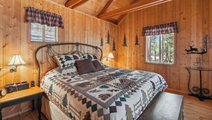 A wooden cabin bedroom with a bed, two windows with plaid curtains, a nightstand with a lamp, and a small table with a decorative item. - Near Trails: Forest Lake Retreat w/ Backyard Oasis (Forest Lakes)