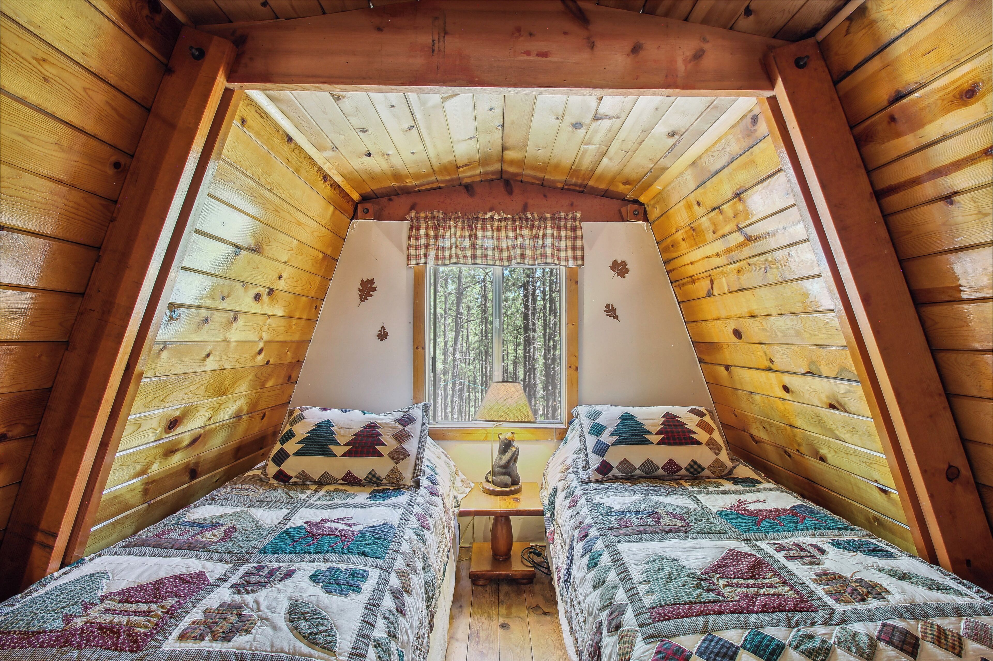 A cozy attic bedroom with wooden walls and a sloped ceiling, featuring two beds with patterned quilts and a small table with a lamp.