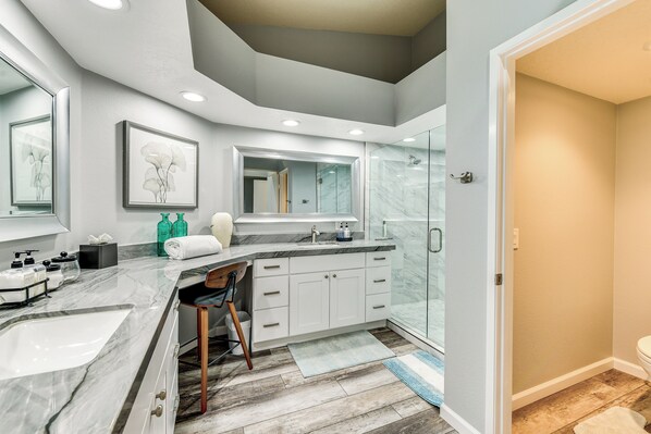 Un baño moderno con tocador de mármol, un espejo grande, cabina de ducha de vidrio y una silla de madera.