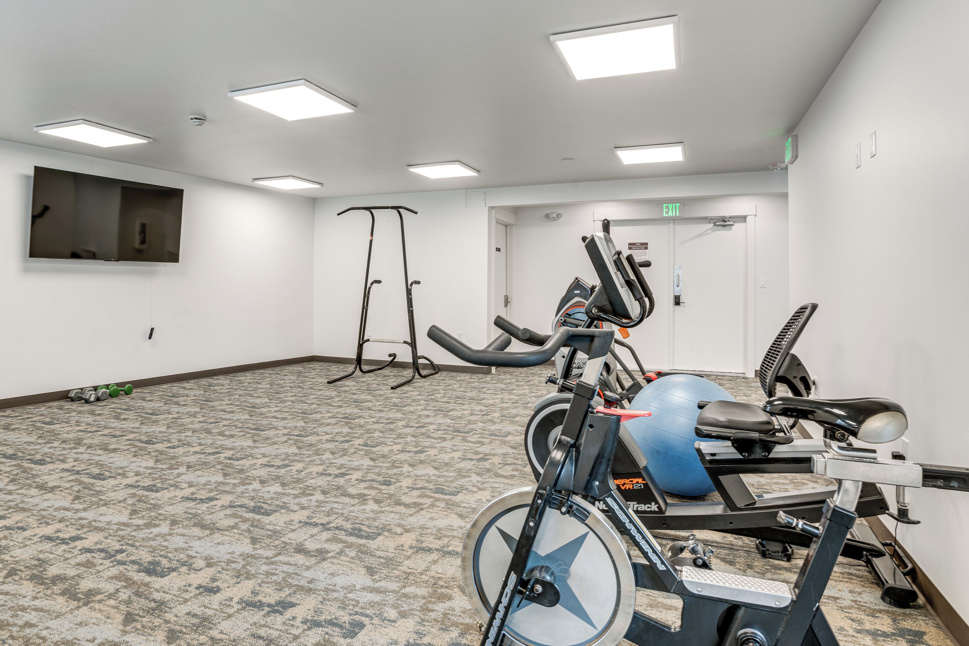 Une salle de sport équipée de matériel d’exercice, avec un téléviseur fixé au mur et une porte de sortie.