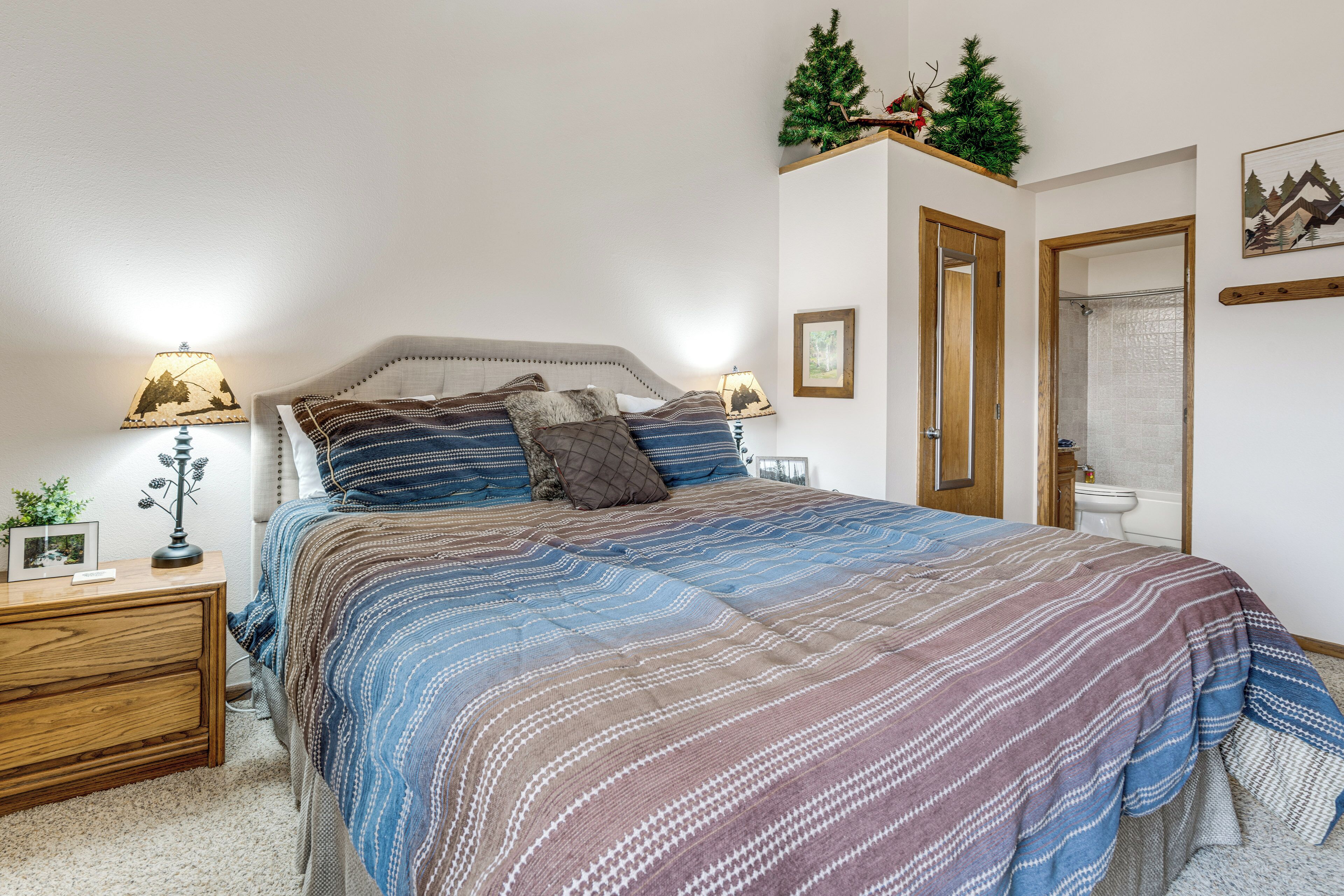 A bedroom with a bed, bedside tables, a nightstand, a mirror, a door, and a small shelf with decorative items.