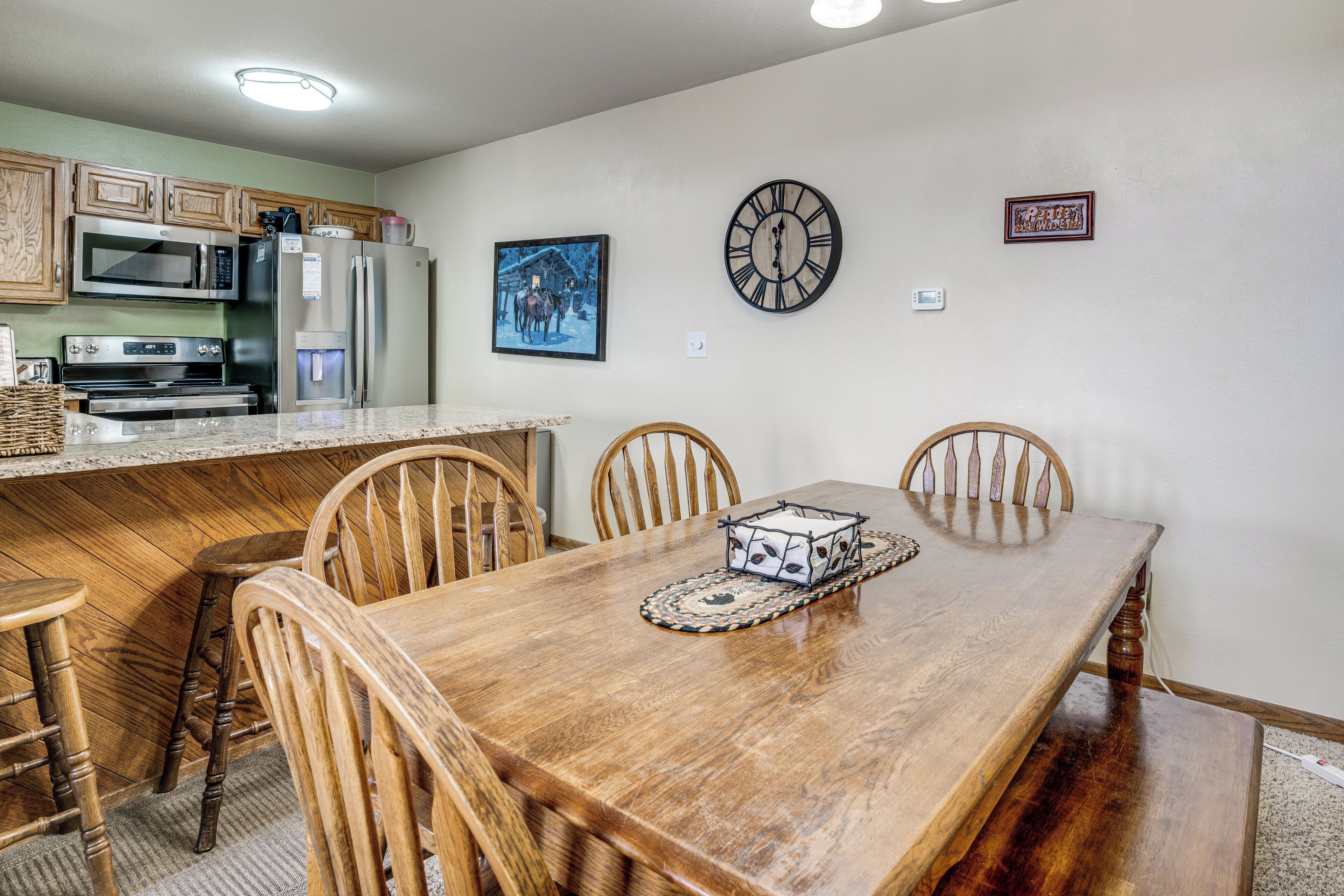 A kitchen with a microwave, refrigerator, and oven, wooden chairs around a dining table, and a clock on the wall.