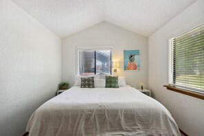 Um quarto com cama, duas janelas com persianas, uma luminária na parede, uma mesinha com uma planta e uma obra de arte com tema de abacaxi na parede.