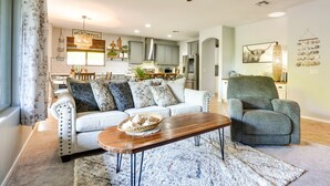A living room with a sofa, a wooden coffee table, and a green armchair. There is a ceiling fan, a chandelier, and a kitchen in the background. - 'sunny V' Arizona Retreat w/ Private Pool & Patio (Surprise)