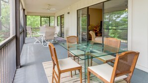 A spacious balcony with a glass table and chairs, a ceiling fan, and a view of trees.