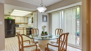 A dining area with a glass table surrounded by wooden chairs, a kitchen in the background, and a large window with blinds.