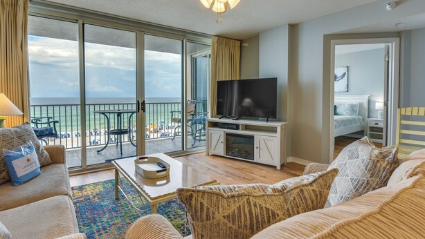 A spacious living room with a large TV, comfortable seating, a balcony with a view of the ocean, and a bedroom with a bed and nightstand. - Fort Walton Beach Rental w/ Pool & Hot Tub Access (Fort Walton Beach)