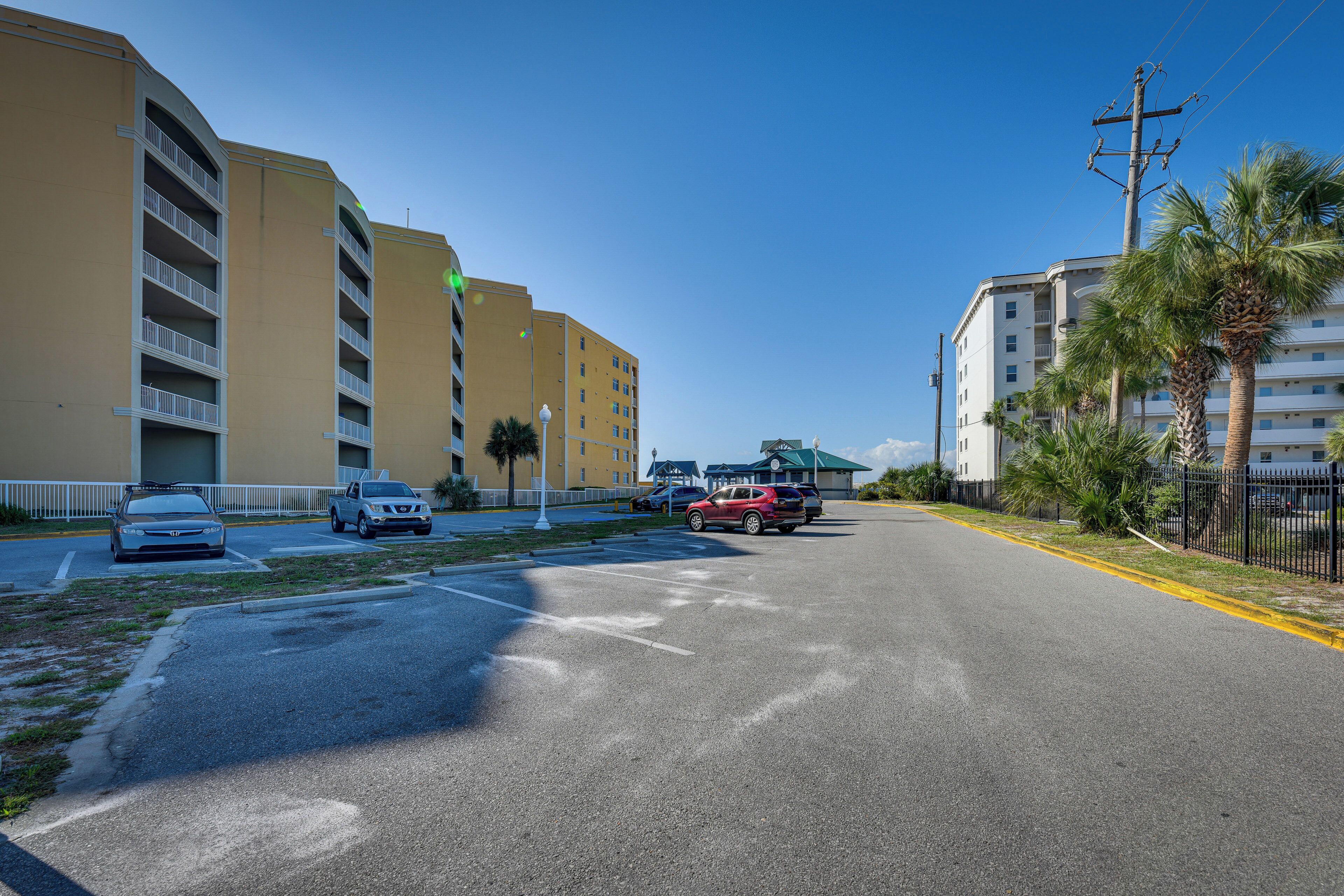 A street with parked cars, a building with balconies, and palm trees.