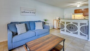 A living room with a blue sofa, a wooden coffee table, and a kitchen area with wooden cabinets and a white refrigerator.