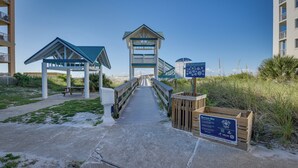 A beachside walkway leading to a beach hut and a lifeguard tower, with a wooden signboard and a wooden crate nearby.