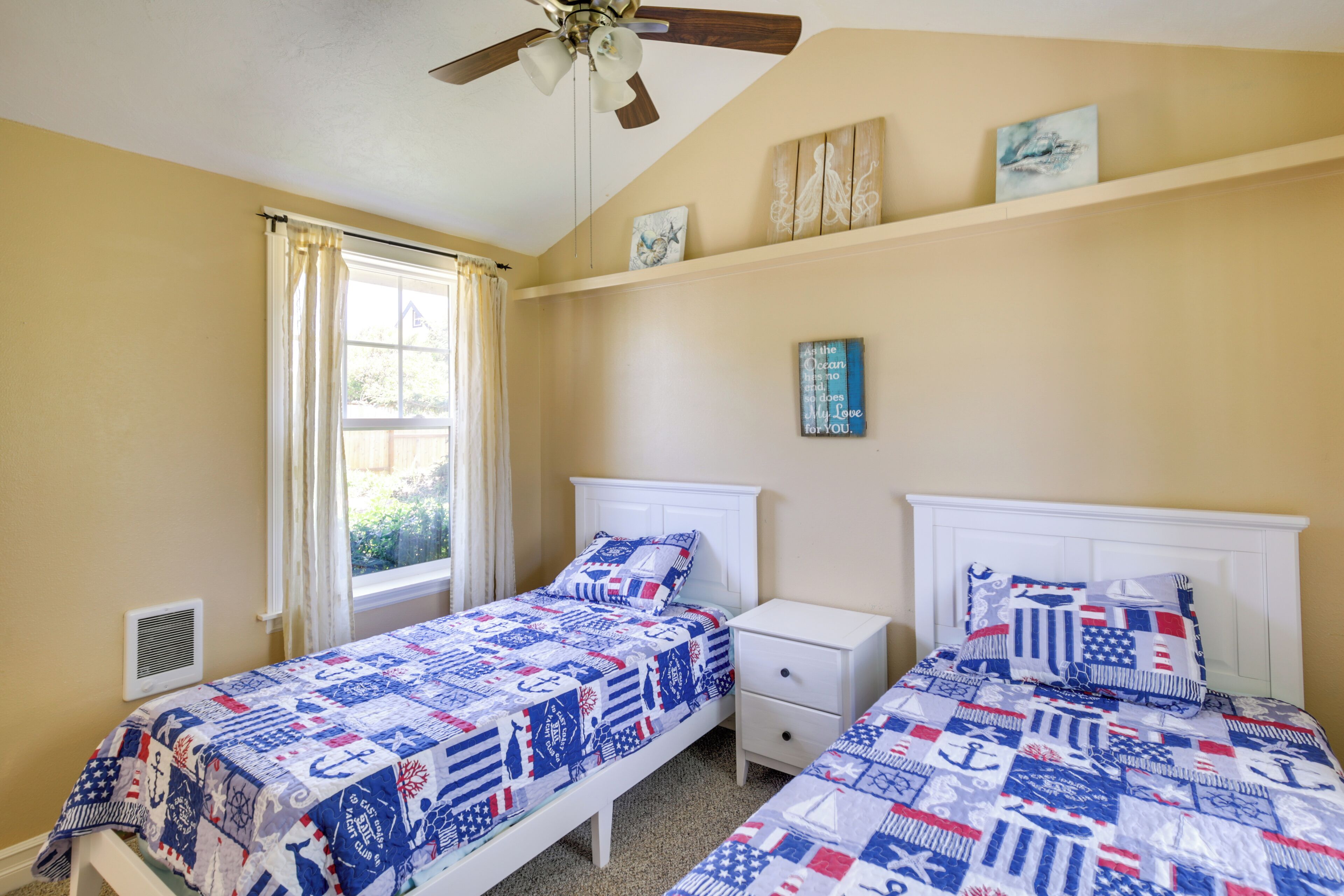 A bedroom with two beds, a ceiling fan, a window with curtains, and a small shelf with artwork.