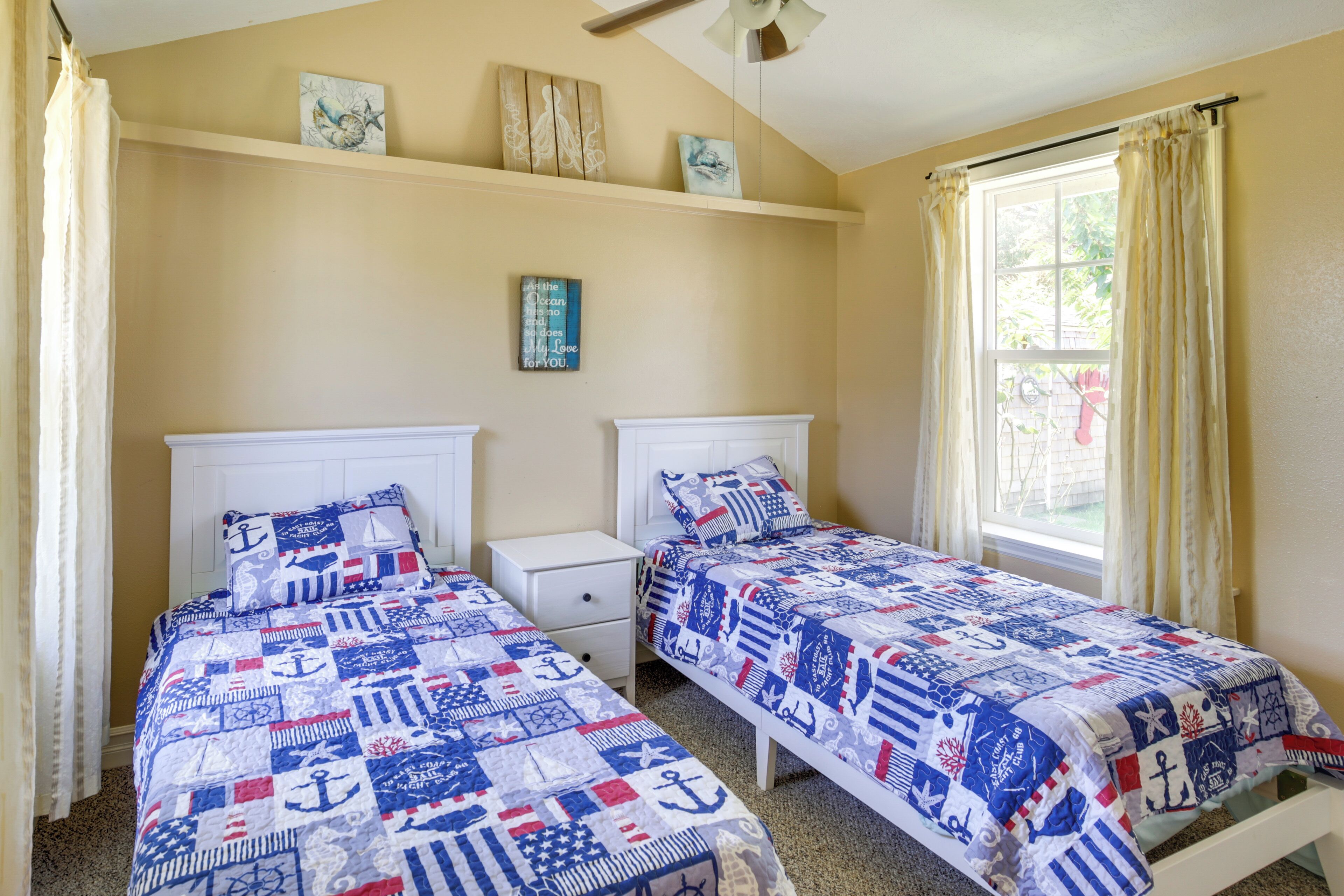 A bedroom with two beds, each with a nautical-themed quilt, a white nightstand, and a window with curtains.