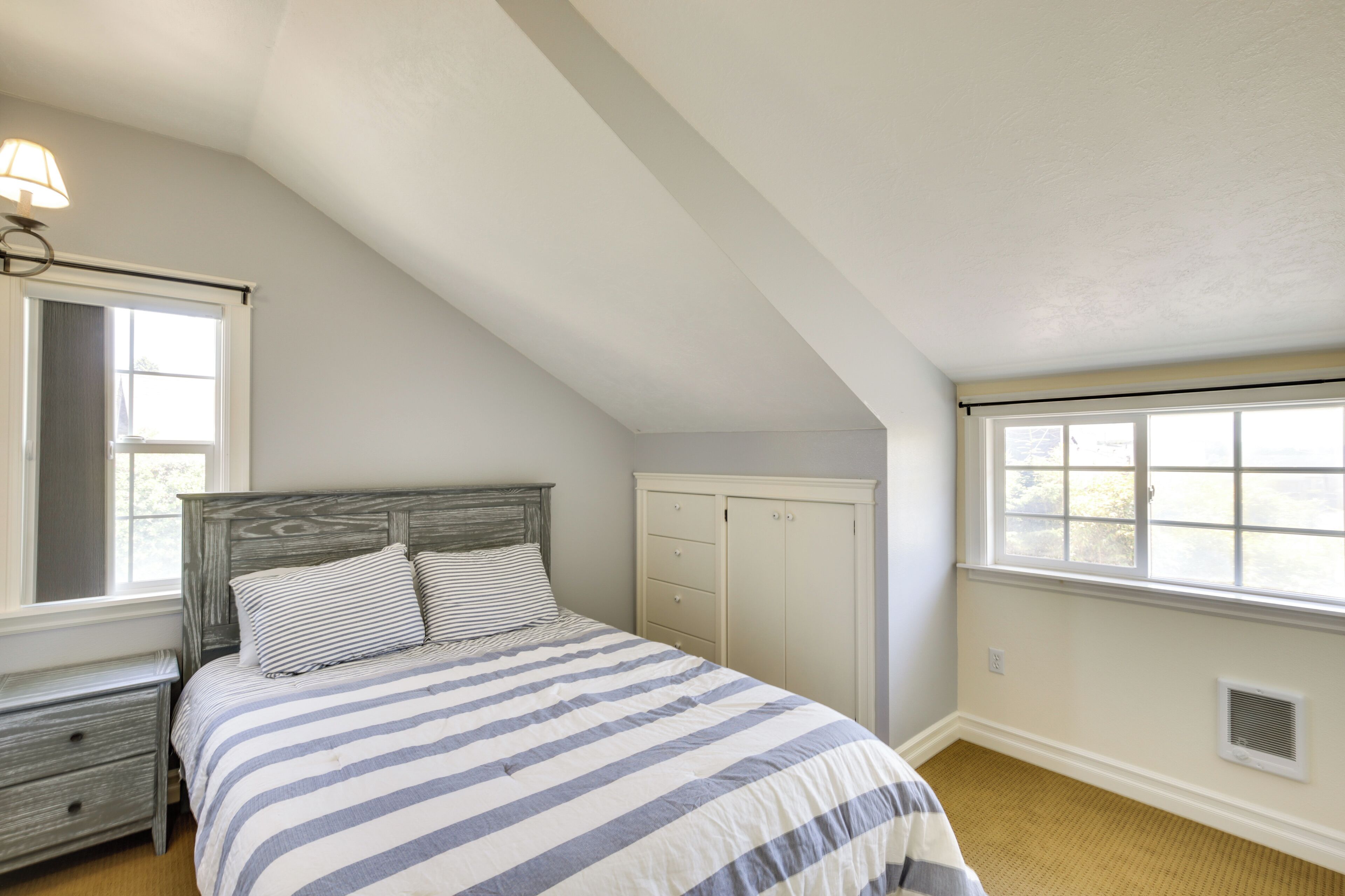Ein Schlafzimmer mit schrägen Wänden, einem Bett mit gestreifter Bettwäsche, einem Nachttisch, einer Kommode und einem Fenster mit Aussicht.