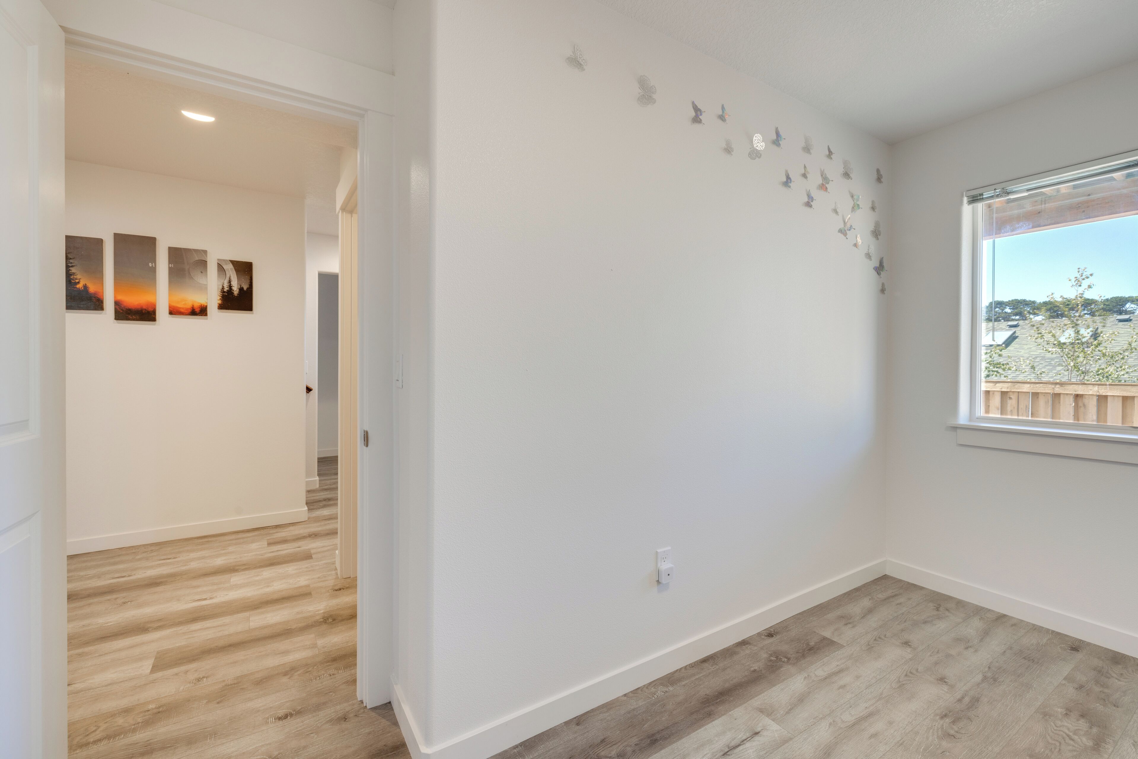 A room with a wooden floor, white walls, a window with a view of the outside, and three wall-mounted pictures.
