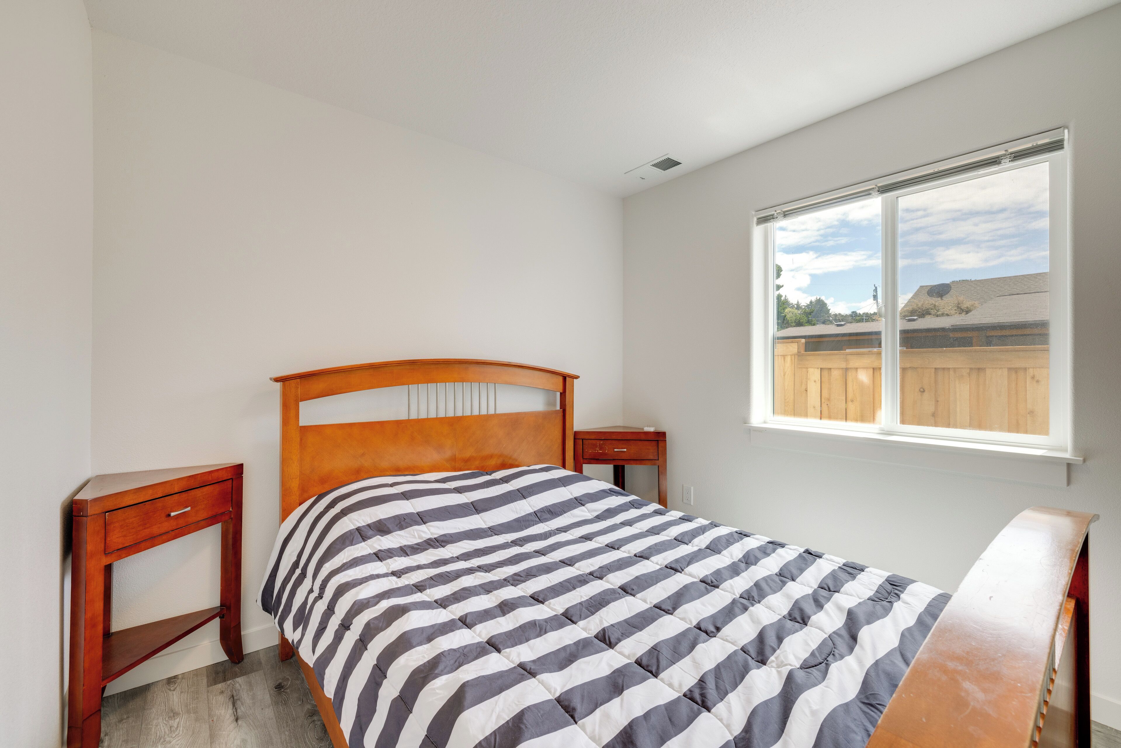 A bedroom with a wooden bed, a nightstand, a window with a view of a wooden fence, and a white wall.