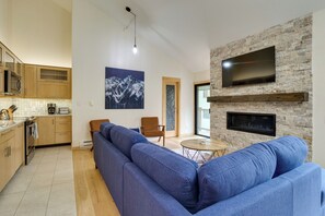 A modern living room with a blue sectional sofa, a fireplace, a flat-screen TV, and a kitchen area with wooden cabinets and a tiled backsplash.