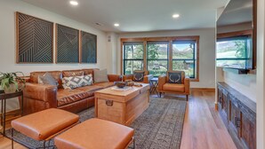 A spacious living room with a leather sofa, ottomans, a wooden coffee table, and a large window offering a view of the outdoors.