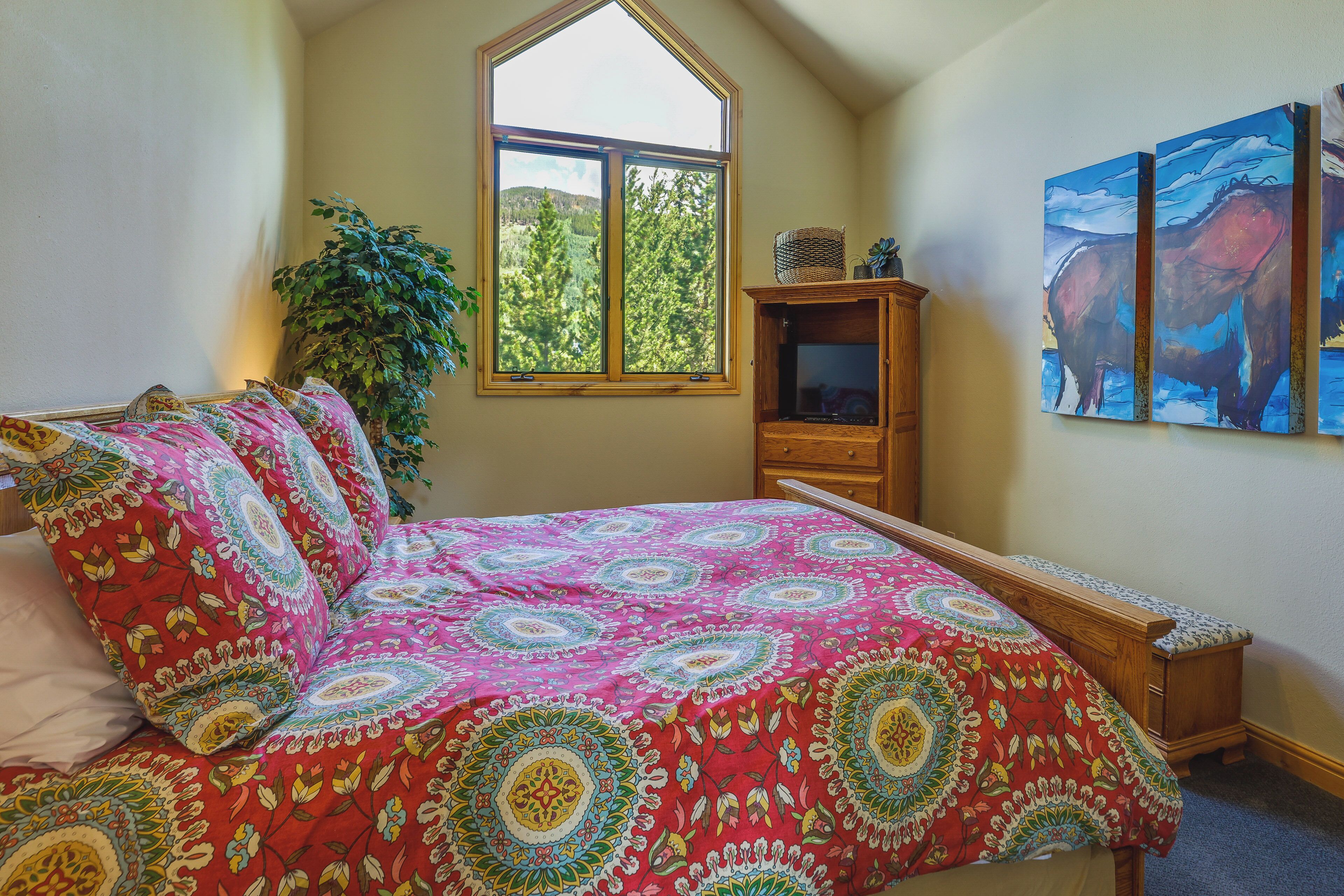 A bedroom with a large window, a bed with a colorful bedspread, a wooden nightstand, and a potted plant.