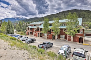 A multi-story building with a red brick facade and green roofs, surrounded by parked cars and trees, with mountains in the background.