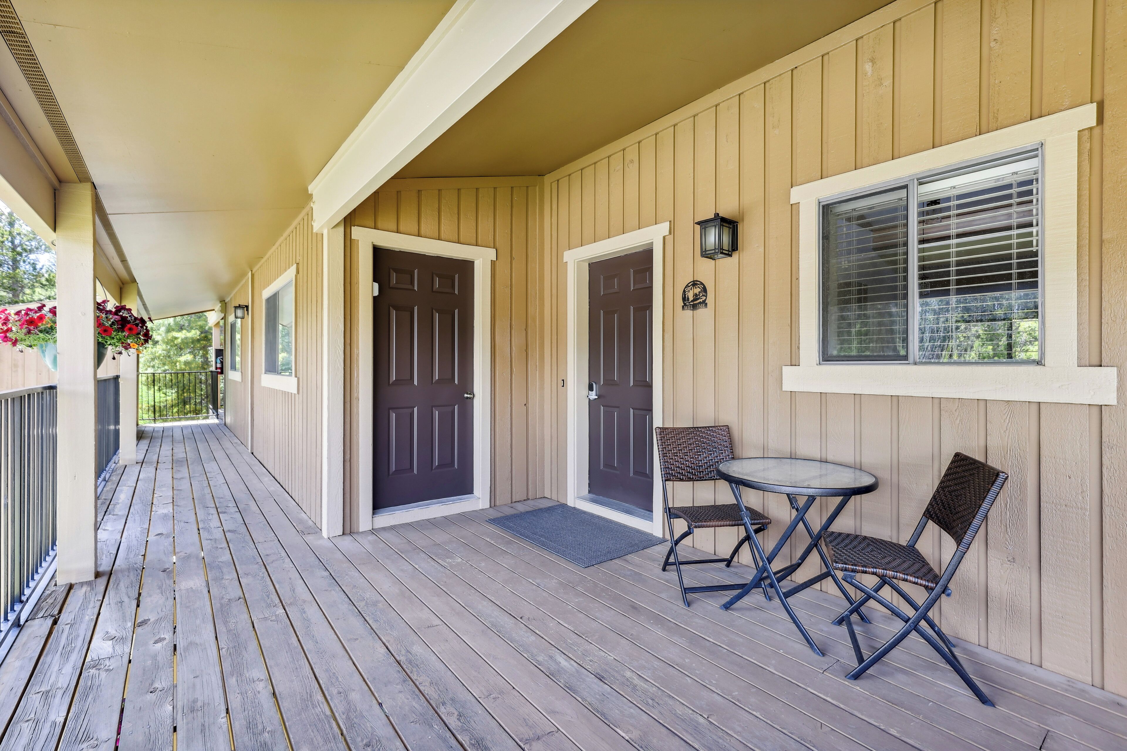 Une véranda couverte avec un plancher en bois, deux portes, une petite table avec des chaises et des jardinières suspendues.