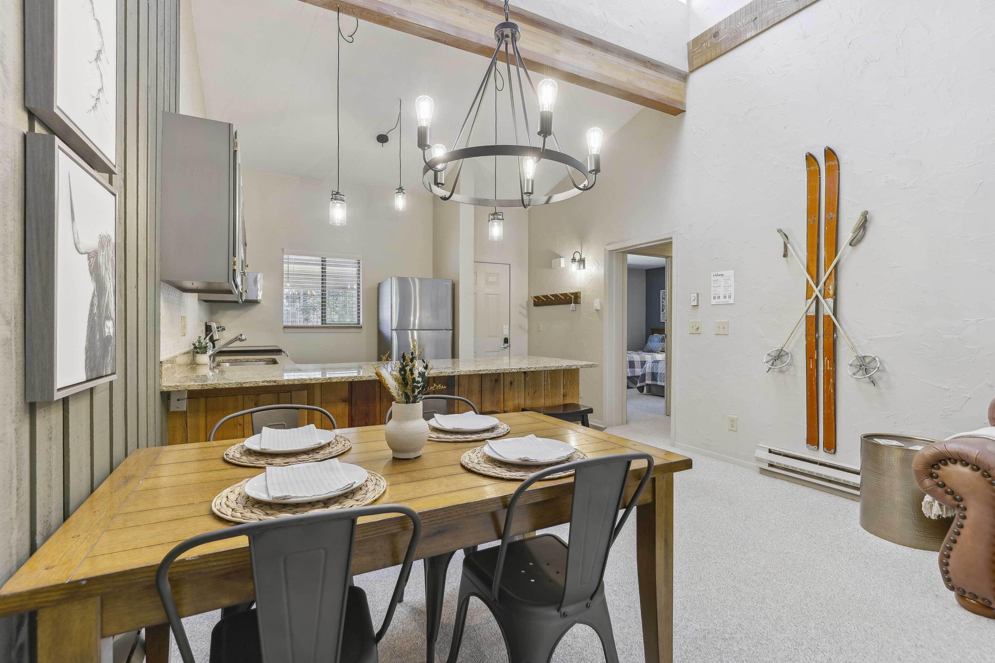 A dining area with a wooden table set for four, a chandelier, and a kitchen in the background.