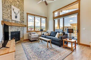 A spacious living room with a stone fireplace, large windows, a cozy sofa, and a wooden coffee table. - Mountain-view Fraser Condo w/ Balcony + Hot Tub! (Fraser)