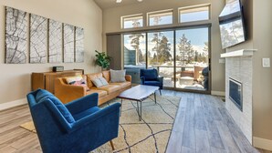 A spacious living room with a large window, a ceiling fan, a fireplace, and a flat-screen TV mounted on the wall. - Fraser Vacation Rental - Shuttle to Winter Park! (Fraser)