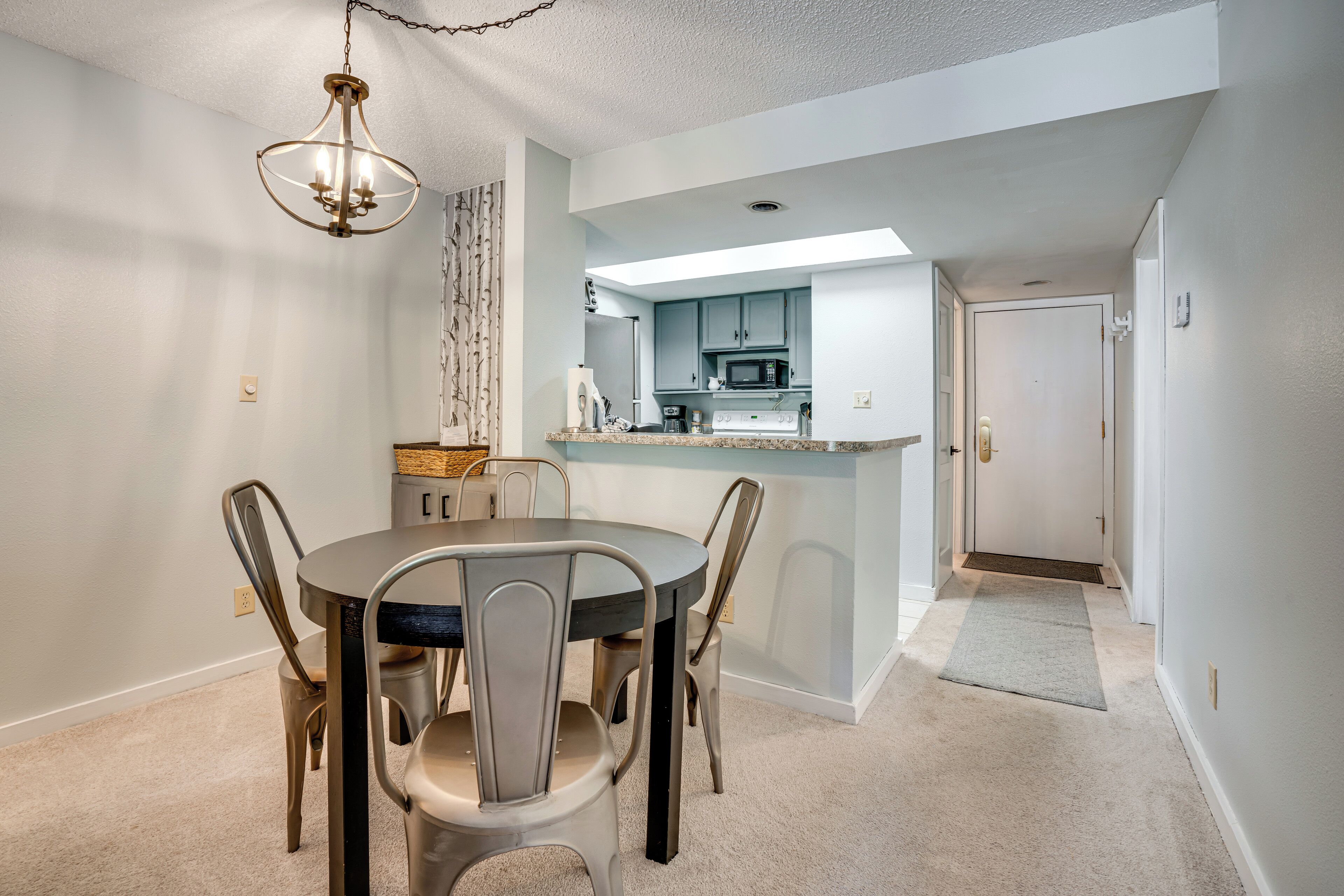 A dining area with a round table and four chairs, a chandelier, and a kitchen in the background.