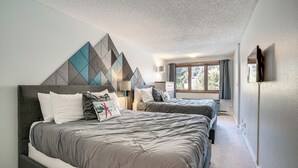 Ein Schlafzimmer mit zwei Betten, einem geometrischen Wanddesign, einem Fenster mit Vorhängen und einem an der Wand montierten Flachbildfernseher.