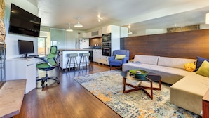 A modern living room with a sofa, coffee table, and a TV. There is a kitchen area with a fridge, oven, and bar stools. - Winter Park Condo w/ Private Balcony: Near Skiing! (Winter Park)