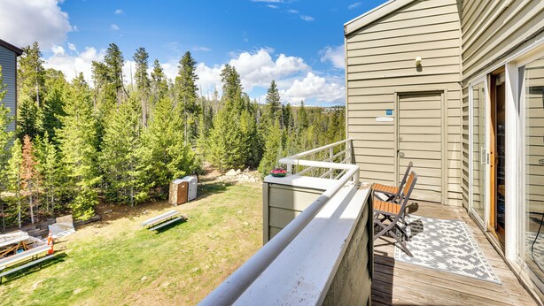 A wooden deck with a view of a forested area and a building with a door and windows. - Winter Park Condo Rental: 4 Mi to Ski Resort! (Winter Park)
