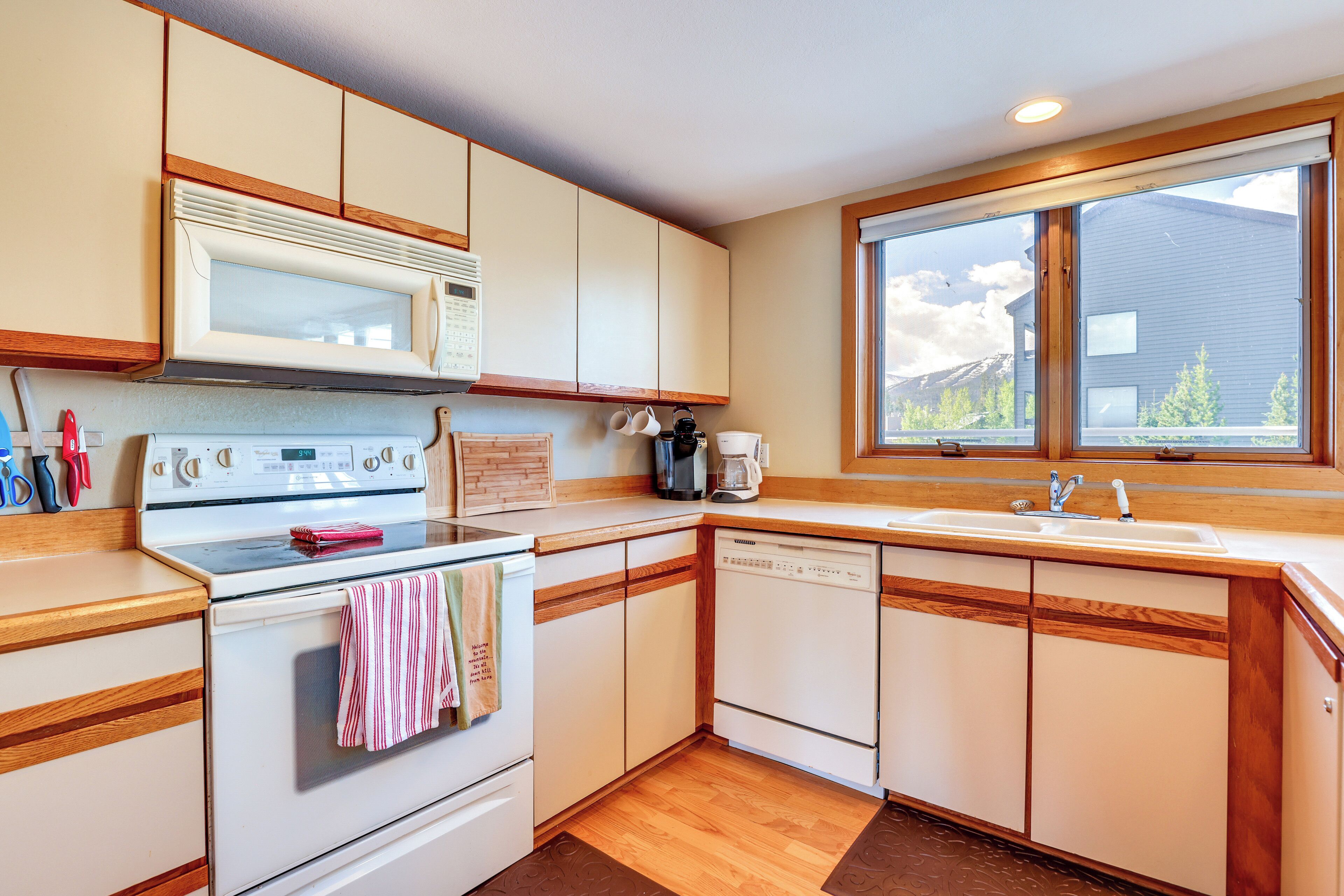 A kitchen with wooden cabinets, a microwave, an oven, a dishwasher, and a window with a view of a house and mountains.