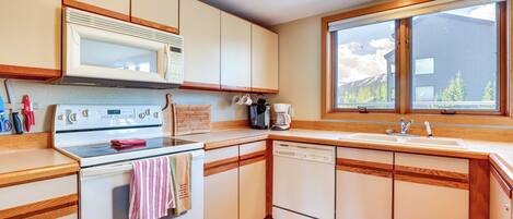 A kitchen with wooden cabinets, a microwave, an oven, a dishwasher, and a window with a view of a house and mountains.