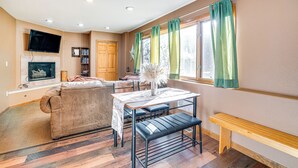 Ein Wohnzimmer mit einer Couch, einem Tisch mit einer Bank, einem Kamin, einem an der Wand montierten Fernseher und einem Fenster mit Vorhängen.
