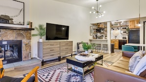 A modern living room with a stone fireplace, a flat-screen TV, a dining area with bar stools, and a kitchen with wooden cabinets. - Pool + Ski-in Access: Copper Mountain Condo! (Copper Mountain)