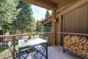 A covered outdoor area with a table and chairs, a stack of firewood, and a view of trees and a building in the background.