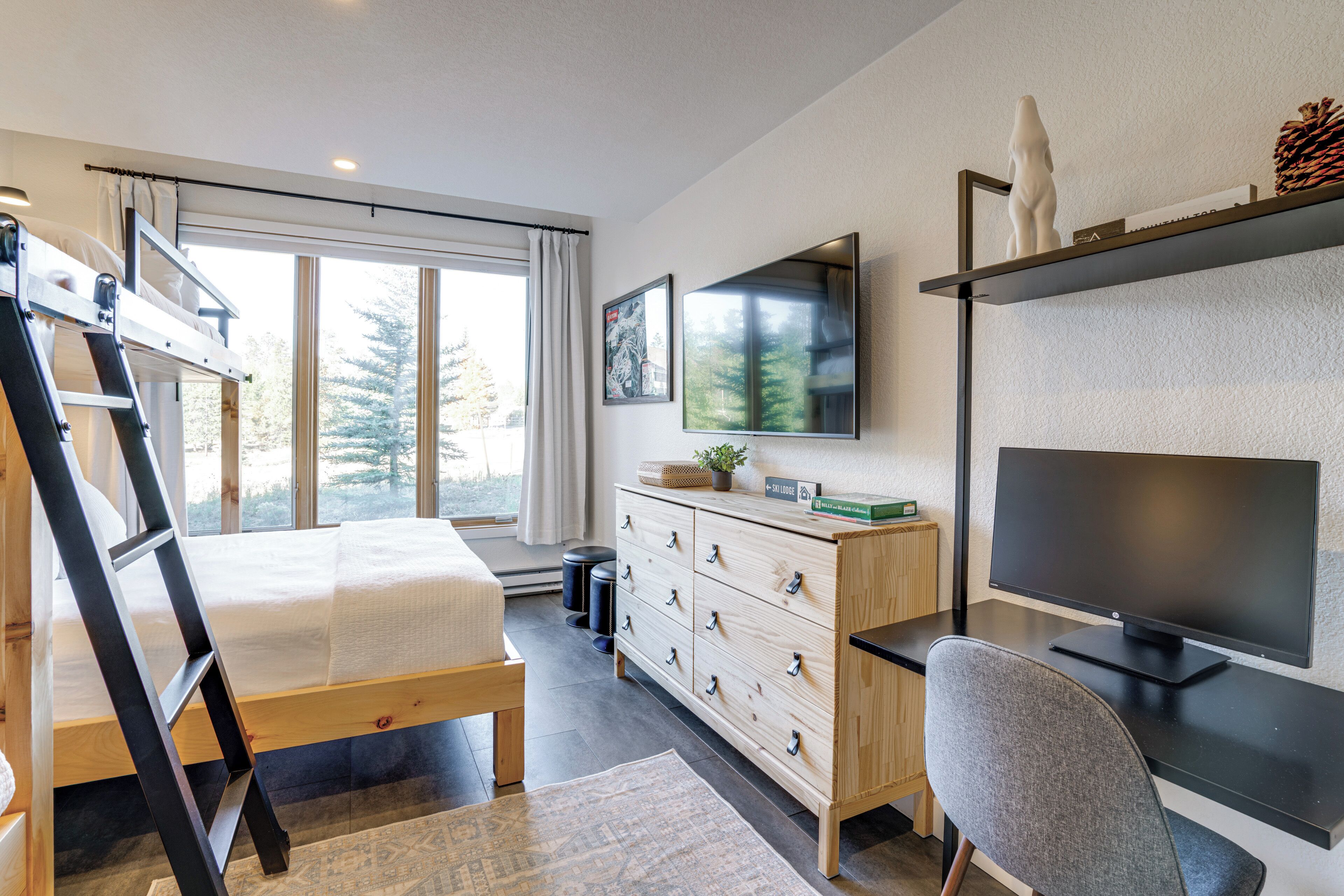 Une chambre moderne avec un lit superposé, un bureau avec un ordinateur, une commode et une grande fenêtre donnant sur les arbres.