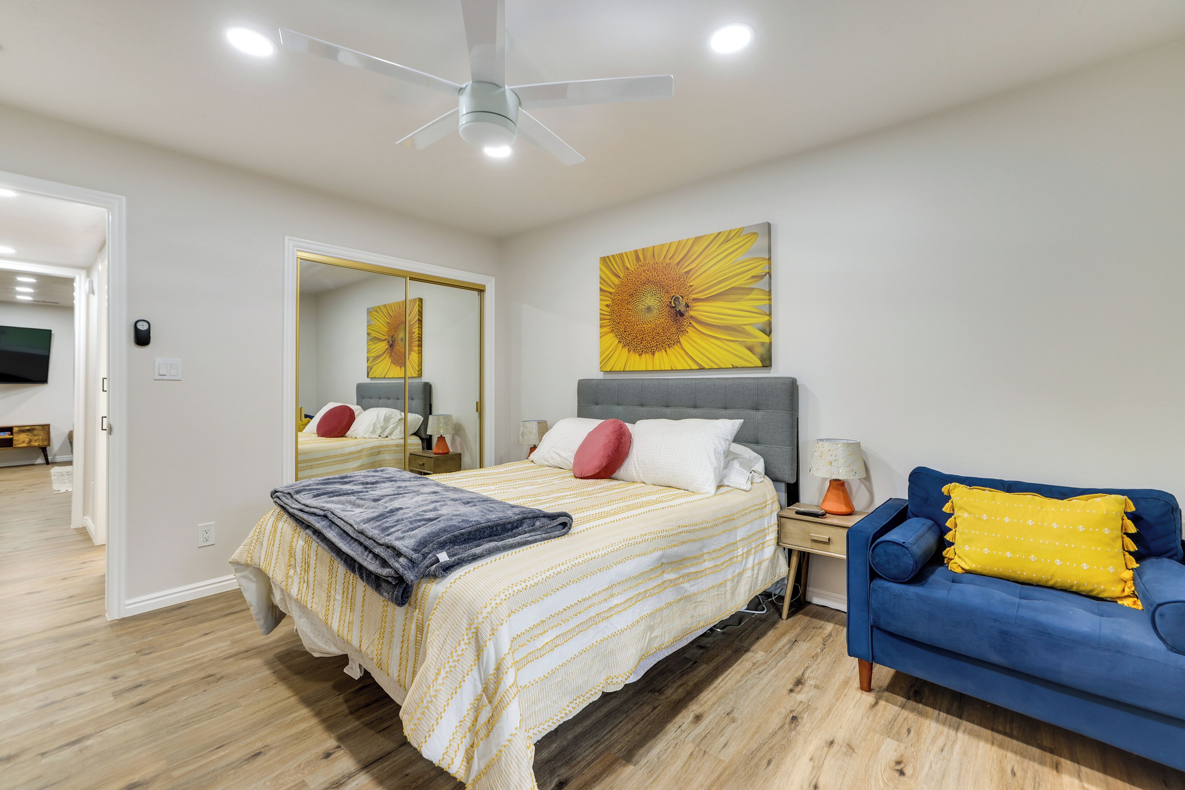 A bedroom with a bed, a nightstand, a lamp, a sofa, a ceiling fan, and a large mirror.