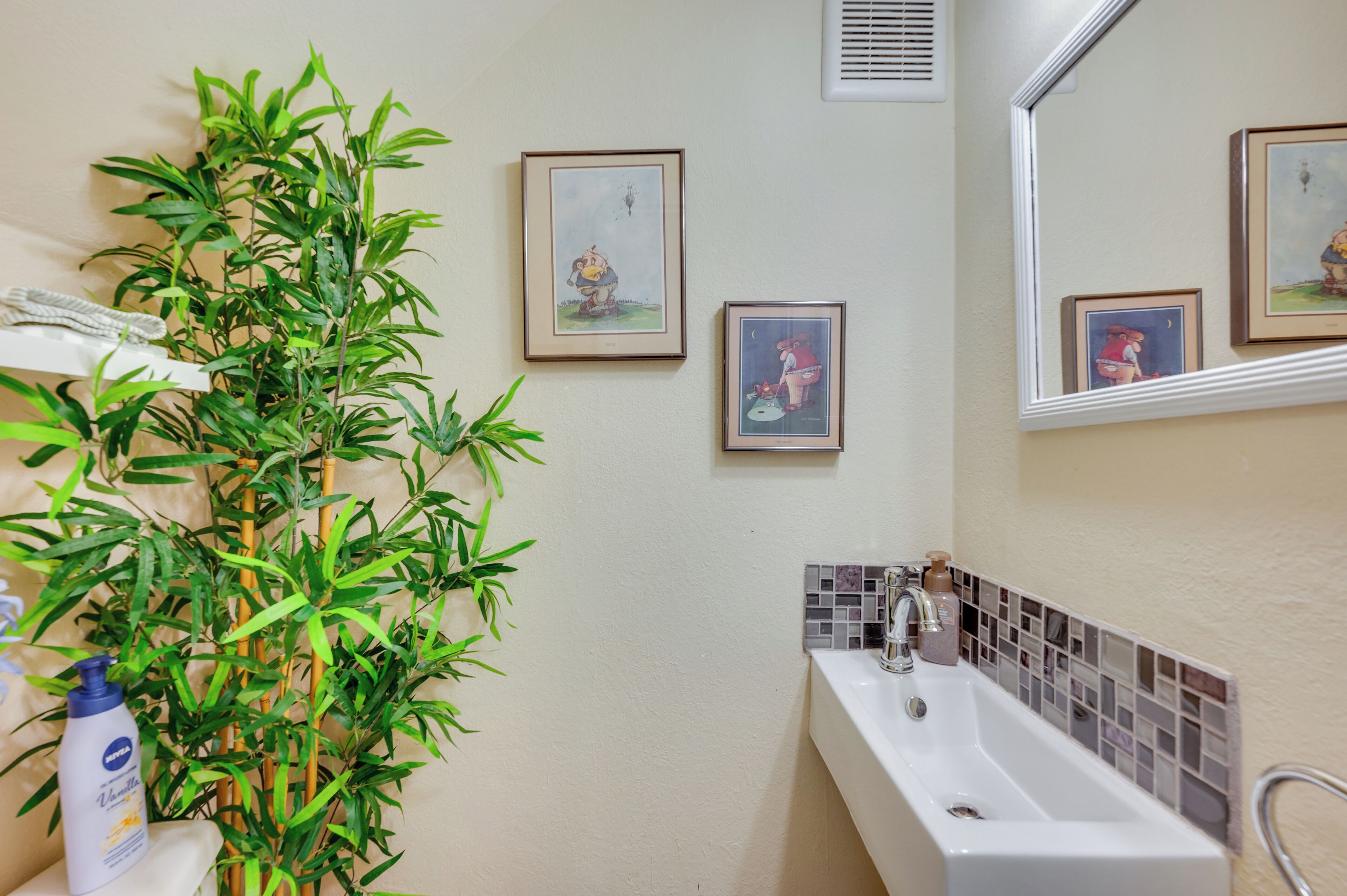 Une salle de bain avec un lavabo, un miroir, des tableaux encadrés au mur et une plante en bambou.