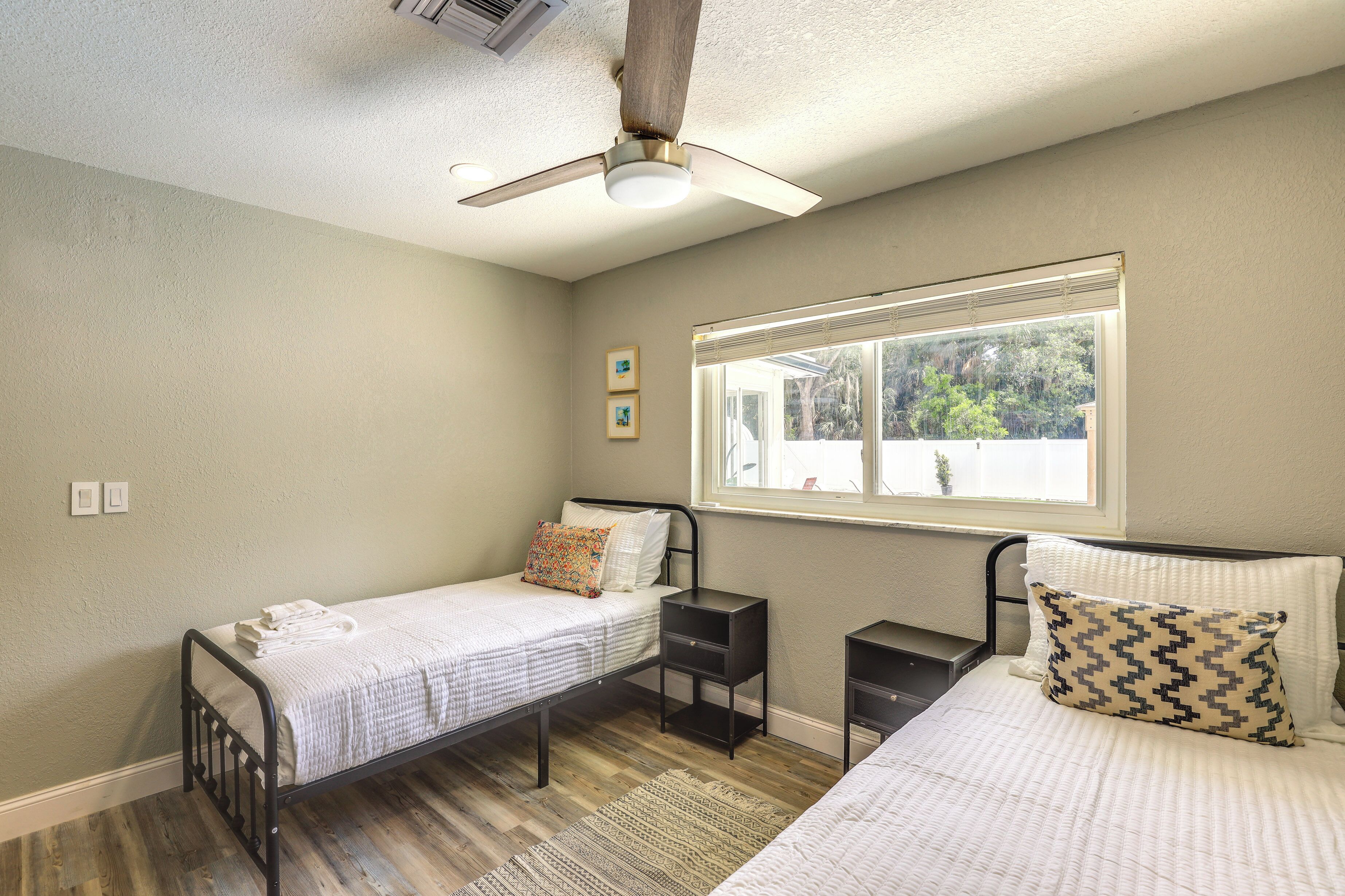 A bedroom with two single beds, a window with blinds, a ceiling fan, and a small bedside table with a lamp.