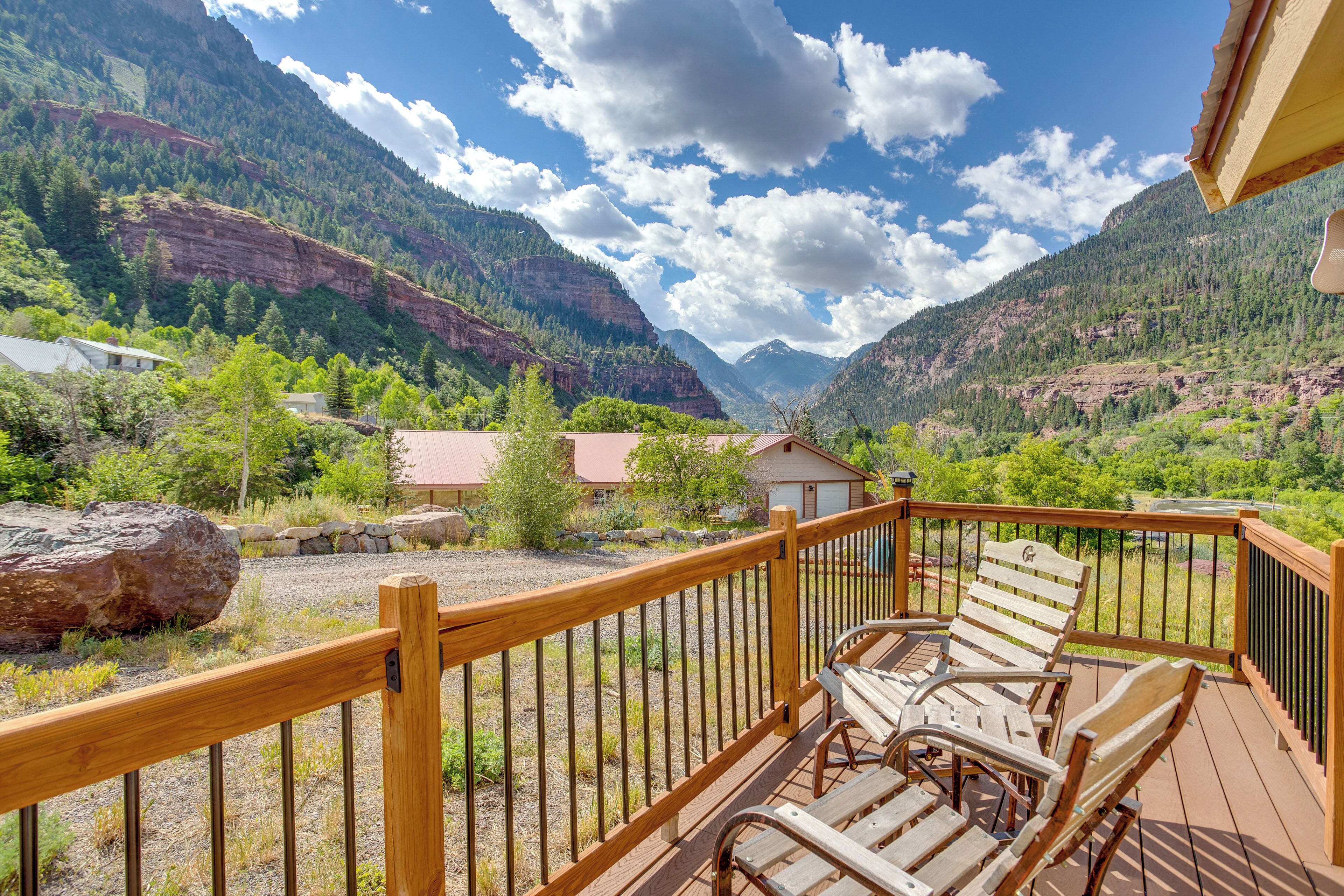 A wooden deck with outdoor seating, overlooking a mountainous landscape with houses and greenery.