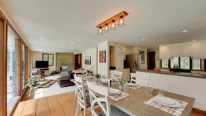 A spacious living area with a wooden dining table set for four, surrounded by white chairs. A television is mounted on the wall, and there is a kitchen area with modern appliances visible in the background. - Mendon Home w/ Hot Tub, Fire Pit & Near Skiing! (Rutland)