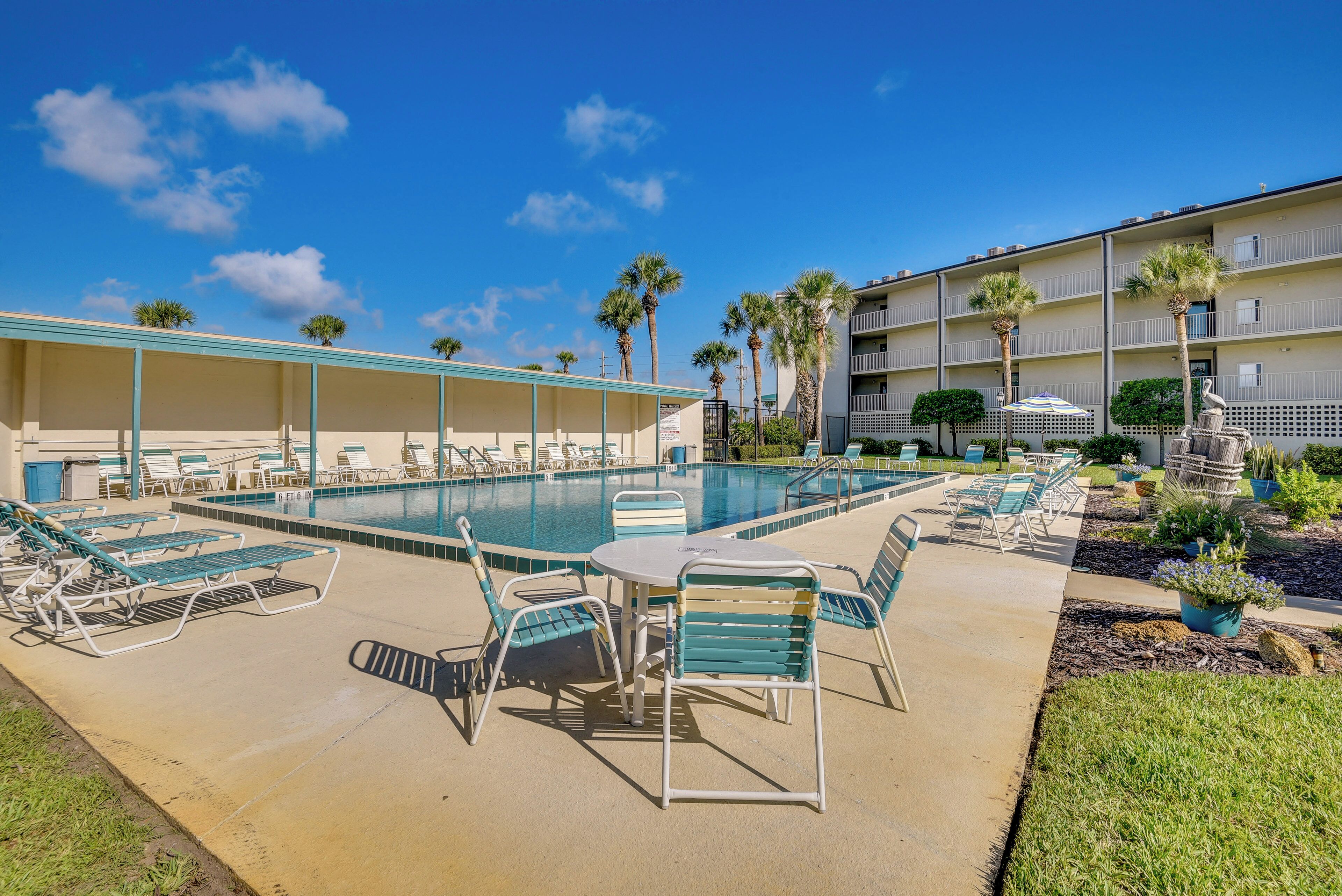 A pool area with lounge chairs and a table, surrounded by a building with balconies and palm trees.